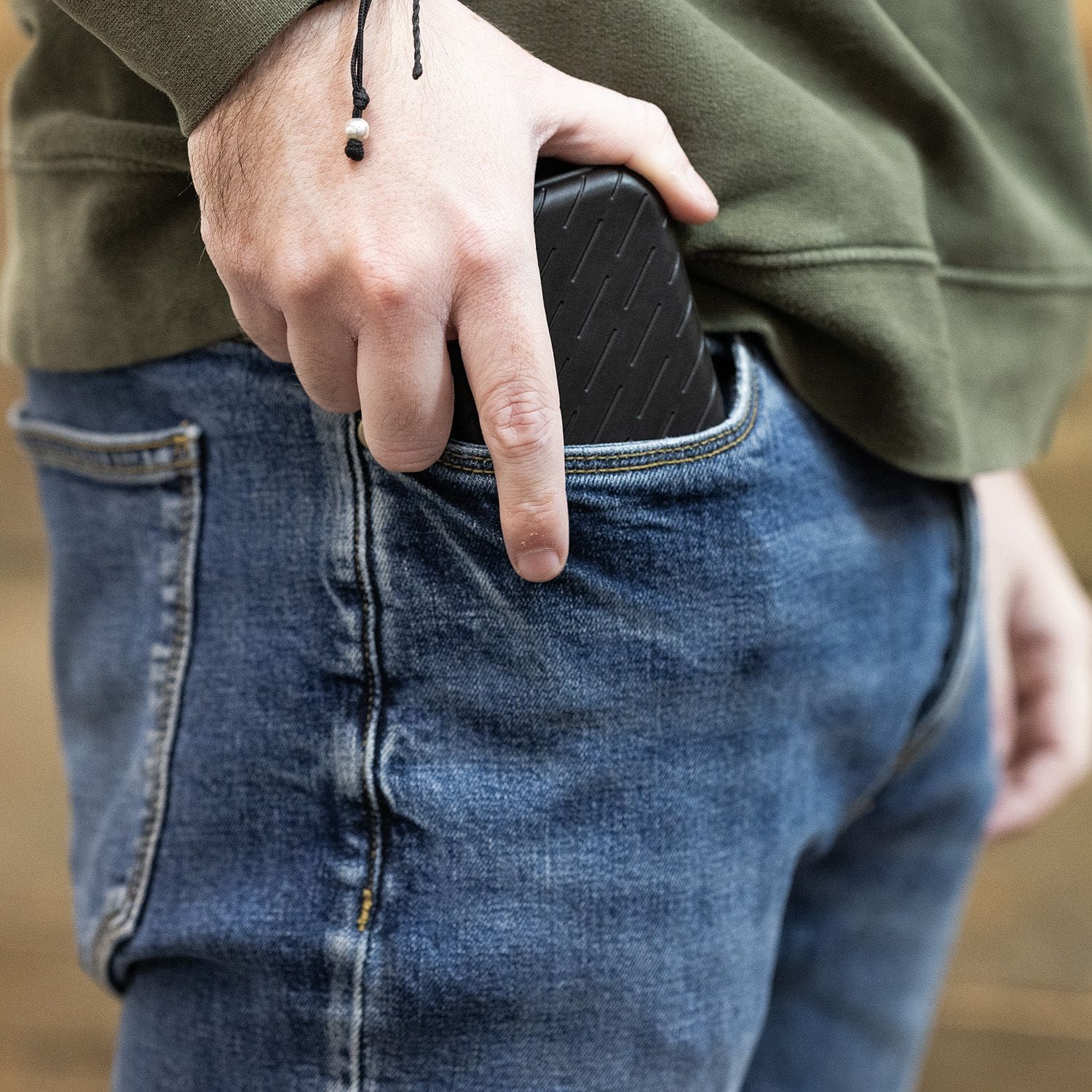 Estuche para dardos Harrows Slimcase Eine Person steckt ein schwarzes Etui in die Hosentasche. Sie trägt eine grüne Jacke und eine blaue Jeans.