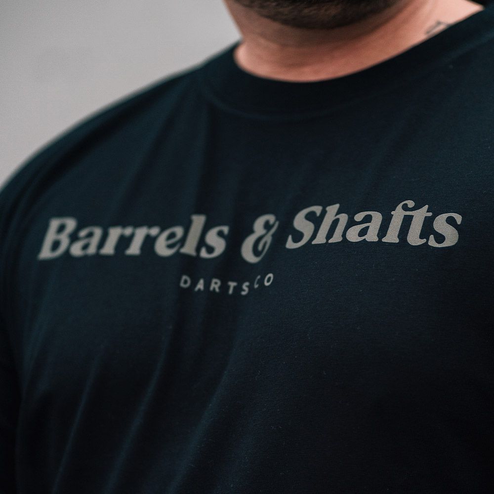 Camiseta Barriles y Ejes - Negro Das Bild zeigt ein schwarzes T-Shirt mit der Aufschrift "Barrels & Shafts". Darunter steht in kleineren Buchstaben "DARTSCO".