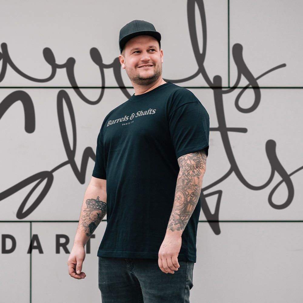 Camiseta Barriles y Ejes - Negro Ein Mann trägt ein schwarzes T-Shirt mit der Aufschrift „Barrels & Shafts“. Er steht vor einer Wand mit passender Schrift im Hintergrund.