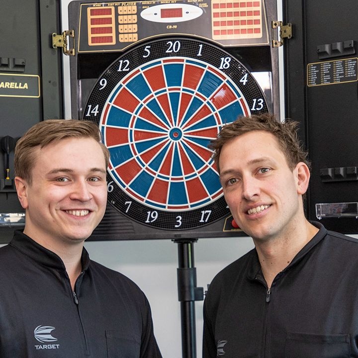 CB90 Zwei Männer stehen lächelnd vor einer elektronischen Dartscheibe. Beide tragen schwarze Jacken mit dem Logo „TARGET“.