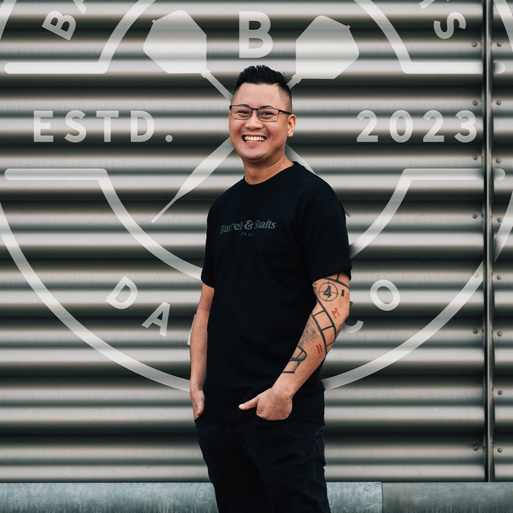 Camiseta Barriles y Ejes - Negro Ein Mann trägt ein schwarzes "Barrels and Shafts"-T-Shirt und steht vor einer metallenen Wand mit großem Logo. Er lächelt und hat markante Tattoos am rechten Arm.