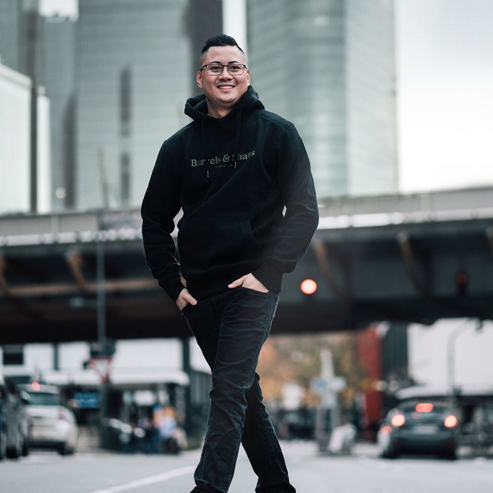 Sudadera con capucha Barrels and Shafts - Negro Eine Person trägt einen schwarzen "Barrels and Shafts" Hoodie und steht entspannt auf einer Straße. Im Hintergrund sind unscharfe Gebäude und Autos zu sehen.