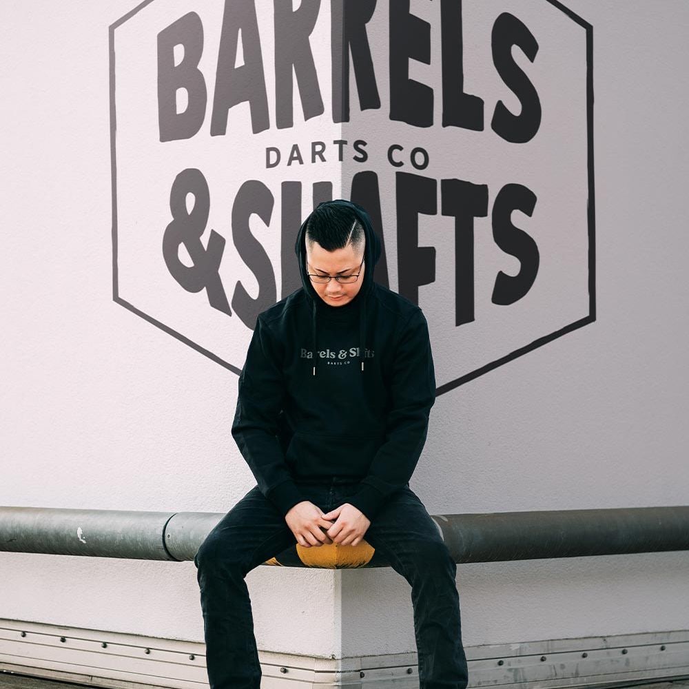 Sudadera con capucha Barrels and Shafts - Negro Eine Person trägt einen schwarzen "Barrels and Shafts" Hoodie und sitzt auf einer Bank. Im Hintergrund ist ein großes Wandlogo mit der Aufschrift "Barrels & Shafts Darts Co" zu sehen.
