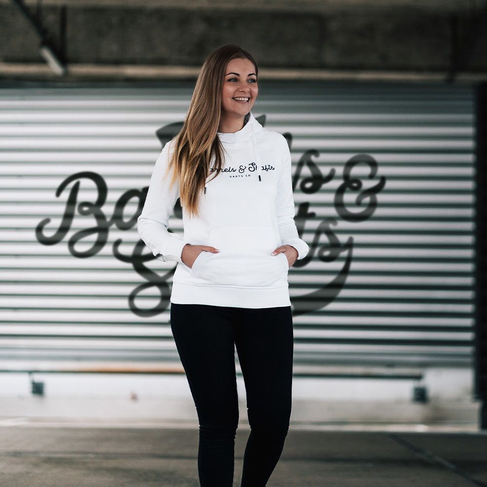Sudadera con capucha Barrels and Shafts - Mujer - Blanco Eine Frau trägt einen weißen "Barrels and Shafts" Damen Hoodie. Sie steht vor einem Hintergrund mit großem Schriftzug und lächelt.