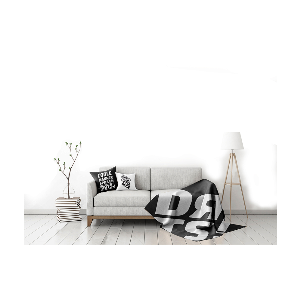 DRTS_K1_DRTS_Kissen_Schwarz_3 Das Bild zeigt ein modern eingerichtetes Wohnzimmer mit einem hellgrauen Sofa. Auf dem Sofa liegt das Produkt "SPORT1 DRTS. Kissen" mit auffälligem schwarzen und weißen Schriftzug.
