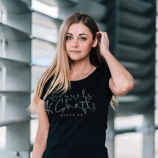 Camiseta Barriles y Ejes - Mujer - Negro Eine Frau trägt ein schwarzes T-Shirt mit der Aufschrift „Barrels and Shafts Darts Co“. Sie steht vor einem modernen, lichtdurchfluteten Hintergrund.