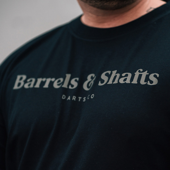 Camiseta Barriles y Ejes - Negro Das Bild zeigt ein schwarzes T-Shirt mit der Aufschrift "Barrels & Shafts". Darunter steht in kleineren Buchstaben "DARTSCO".