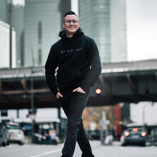 Sudadera con capucha Barrels and Shafts - Negro Eine Person trägt einen schwarzen "Barrels and Shafts" Hoodie und steht entspannt auf einer Straße. Im Hintergrund sind unscharfe Gebäude und Autos zu sehen.