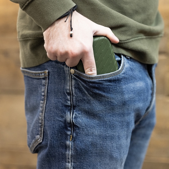 Estuche para dardos Harrows Slimcase Eine Person steckt ein grünes Etui in die vordere Hosentasche einer Jeans. Das Etui hat eine strukturierte Oberfläche und passt gut in die Tasche.