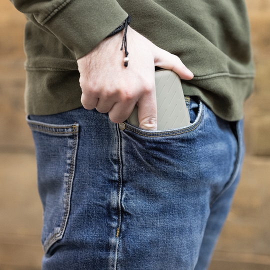 Estuche para dardos Harrows Slimcase Eine Person steckt eine graue Darttasche in ihre Hosentasche. Die Person trägt eine blaue Jeans und einen olivgrünen Pullover.