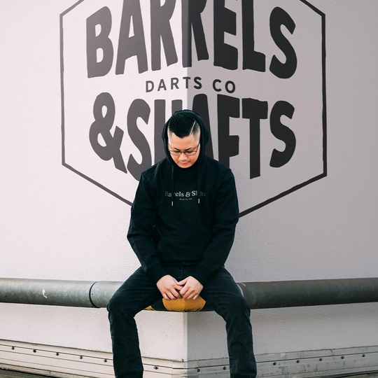 Sudadera con capucha Barrels and Shafts - Negro Eine Person trägt einen schwarzen "Barrels and Shafts" Hoodie und sitzt auf einer Bank. Im Hintergrund ist ein großes Wandlogo mit der Aufschrift "Barrels & Shafts Darts Co" zu sehen.